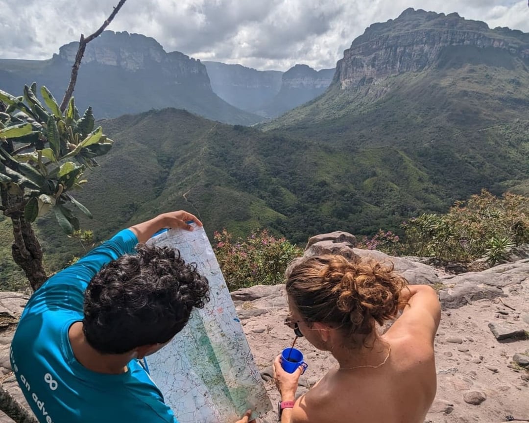 A PlanetaEXO local guide wearing a blue shirt shows a trail map to a female hiker, with a vast green valley and mountains in the background. A PlanetaEXO local guide wearing a blue shirt shows a trail map to a female hiker, with a vast green valley and mountains in the background.