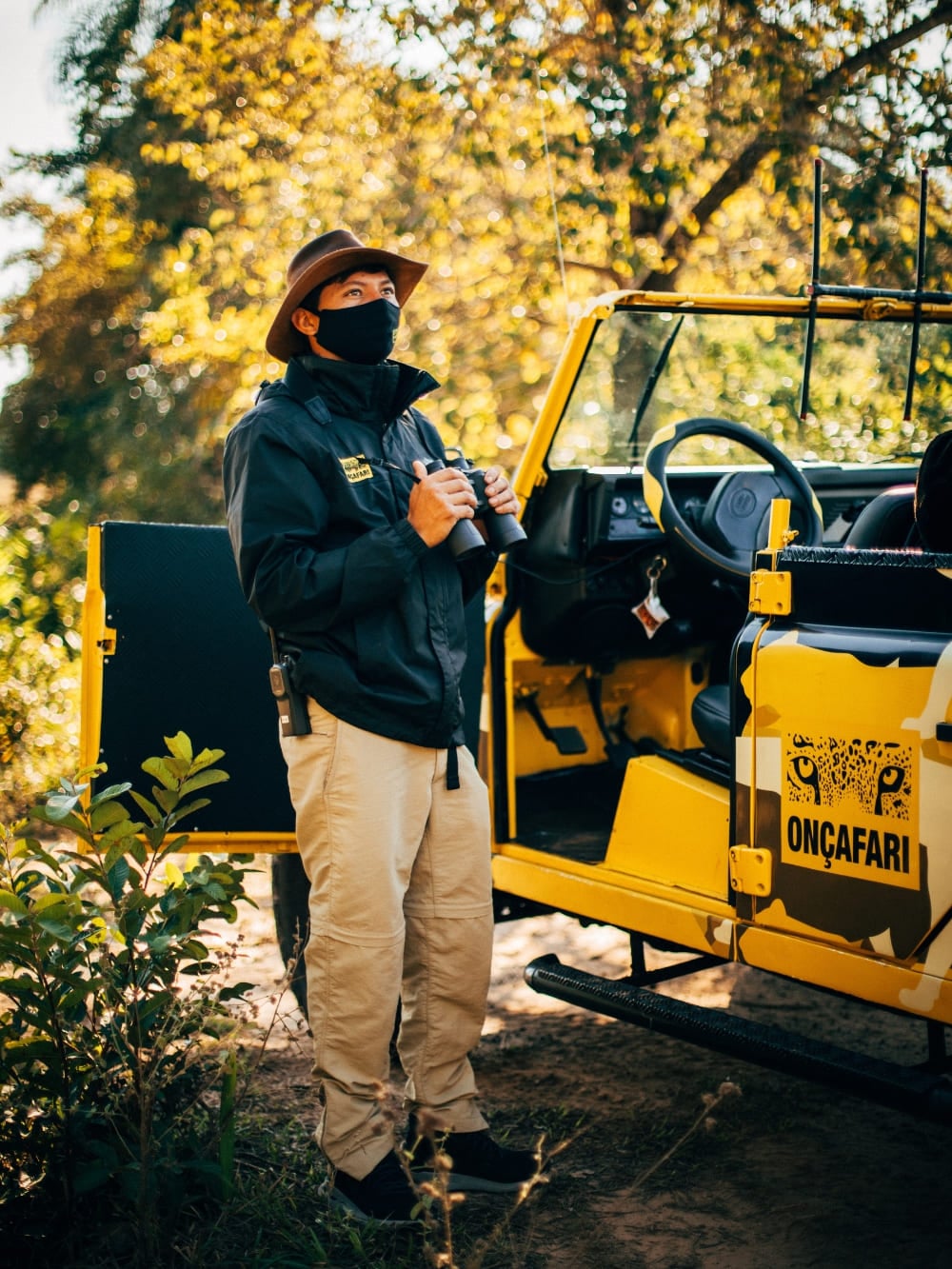 An ecotourism guide holding binoculars standing next to an Onçafari project vehicle. An ecotourism guide holding binoculars standing next to an Onçafari project vehicle.
