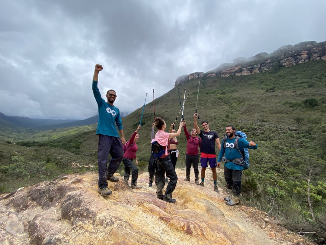 A group of hikers and a PlanetaEXO guide celebrate with raised arms on a rocky peak, surrounded by green mountains under a cloudy sky. A group of hikers and a PlanetaEXO guide celebrate with raised arms on a rocky peak, surrounded by green mountains under a cloudy sky.