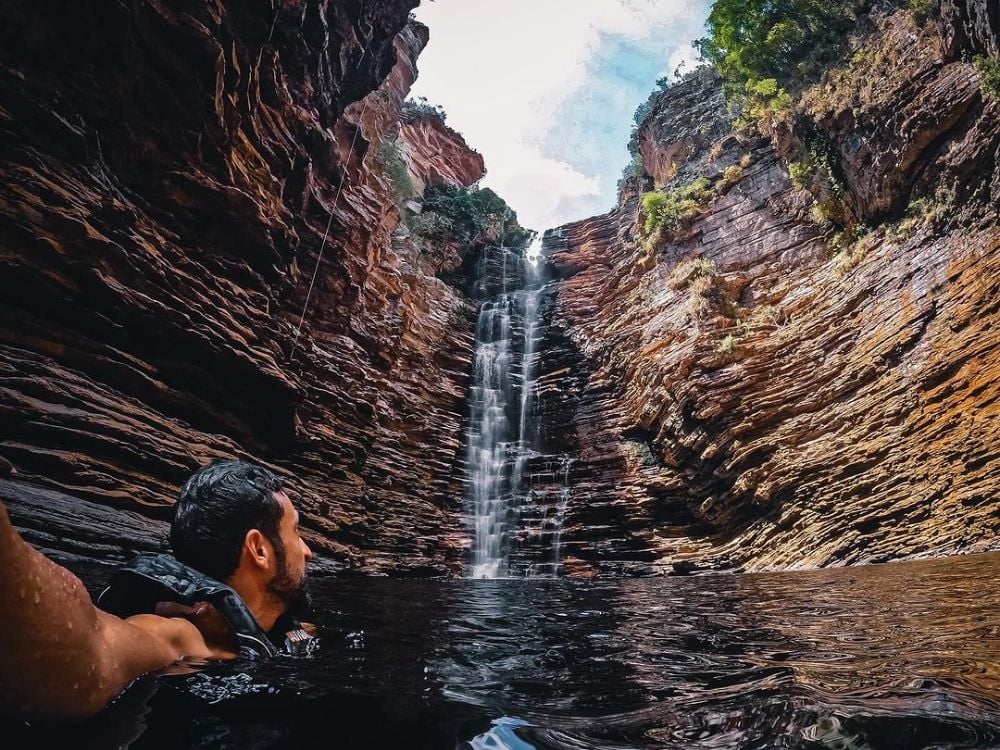 A visitor swims in a dark pool beneath a cascading waterfall surrounded by towering layered rock formations in Chapada Diamantina. A visitor swims in a dark pool beneath a cascading waterfall surrounded by towering layered rock formations in Chapada Diamantina.
