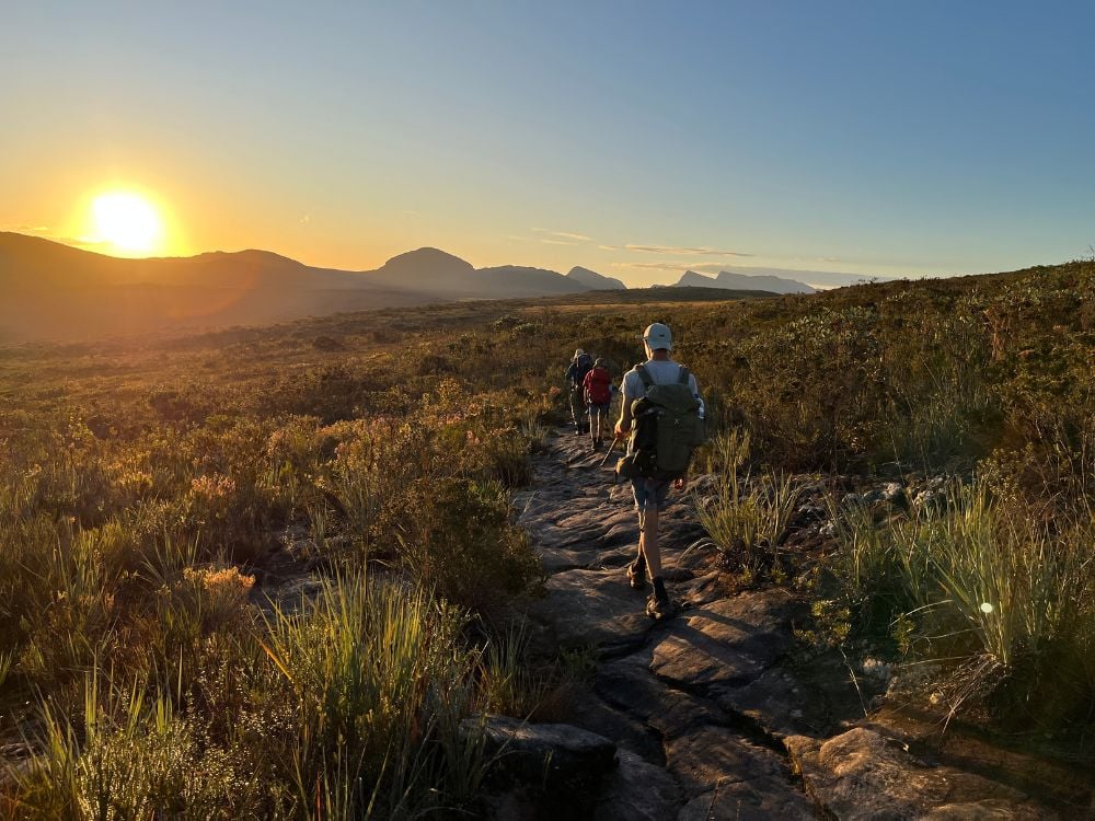 A group of hikers with backpacks walks along a rocky trail towards the mountains during a bright sunset in Chapada Diamantina. A group of hikers with backpacks walks along a rocky trail towards the mountains during a bright sunset in Chapada Diamantina.