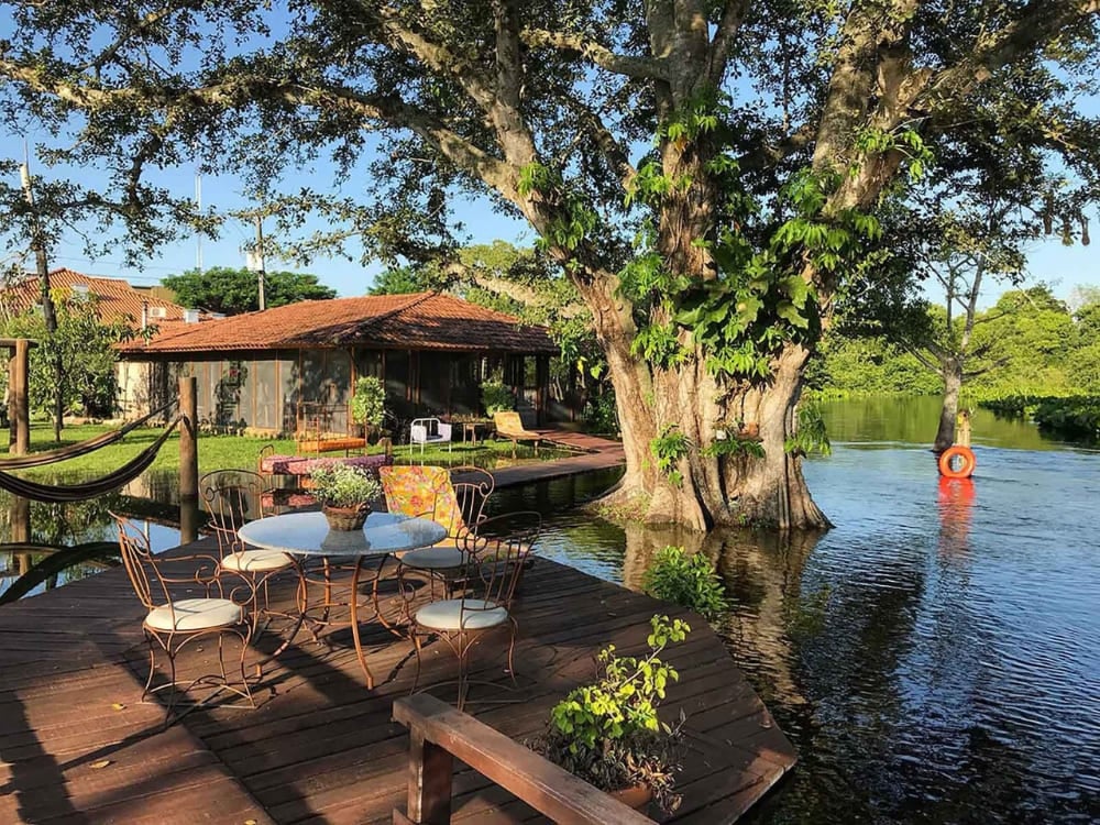 A wooden deck with a table and chairs sits beside a tranquil lake and a massive tree at Refúgio da Ilha ecolodge. A wooden deck with a table and chairs sits beside a tranquil lake and a massive tree at Refúgio da Ilha ecolodge.