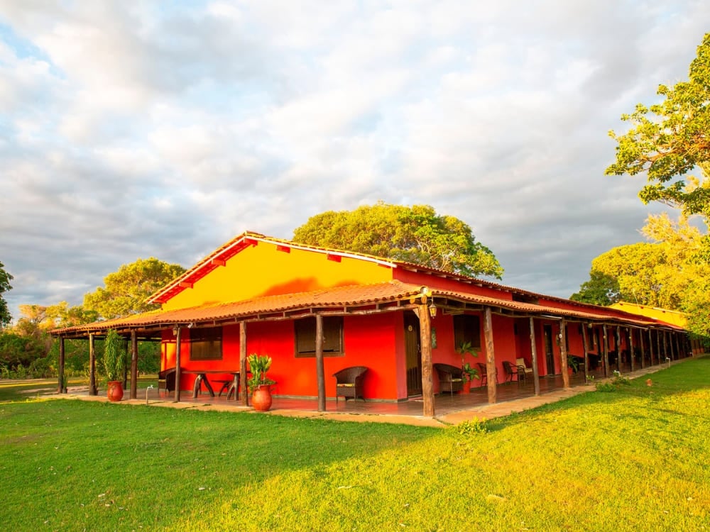 The red exterior of the SouthWild Pantanal Lodge features a shaded wooden porch for guests seeking an authentic stay. The red exterior of the SouthWild Pantanal Lodge features a shaded wooden porch for guests seeking an authentic stay.