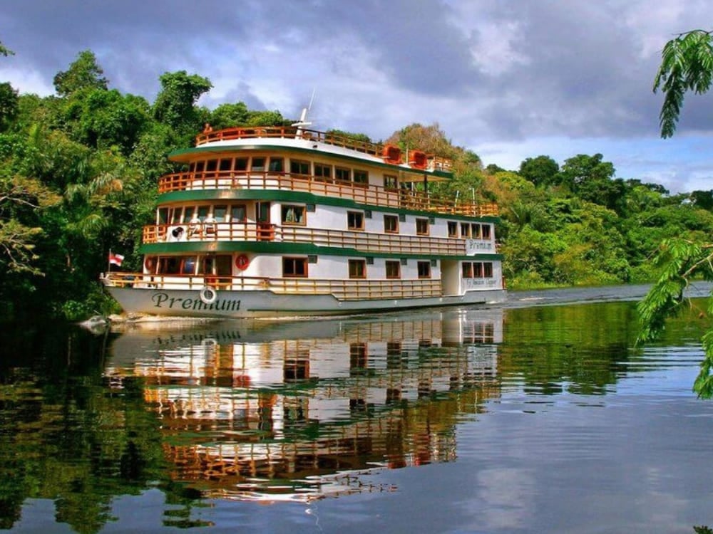 The Amazon Clipper Premium riverboat cruising along the Amazon River, reflecting in the calm water against a backdrop of dense green rainforest. The Amazon Clipper Premium riverboat cruising along the Amazon River, reflecting in the calm water against a backdrop of dense green rainforest.