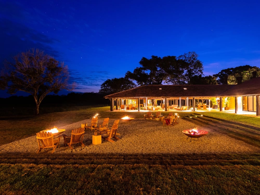 The exterior lounge of Casa Caiman at night, featuring fire pits and wooden chairs under a deep blue starry sky. The exterior lounge of Casa Caiman at night, featuring fire pits and wooden chairs under a deep blue starry sky.