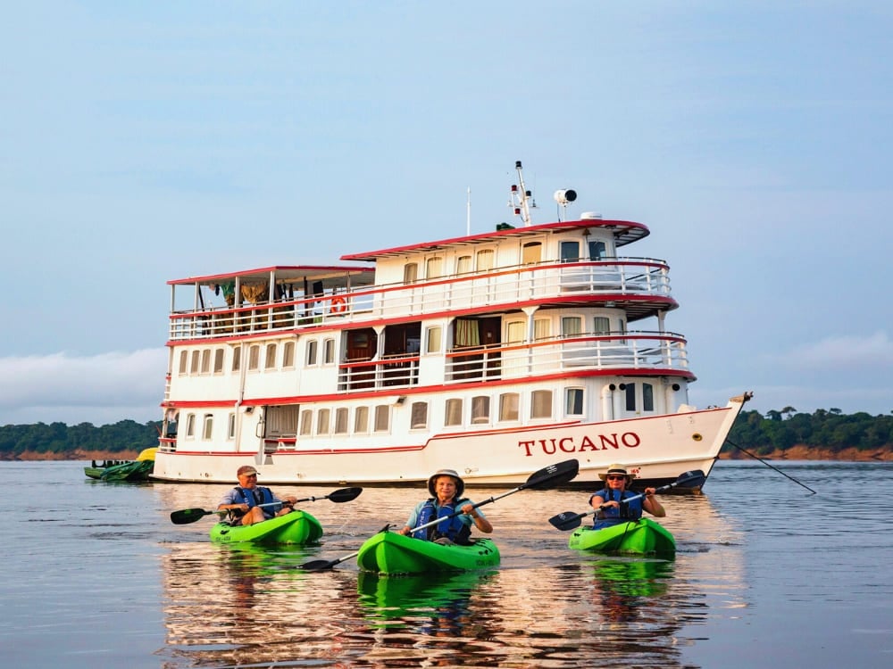 The Tucano motor yacht sailing on the dark waters of the Amazon, designed to explore deep parts of the Central Amazon Conservation Complex. Three people paddle green kayaks on a calm river in front of a multi-deck riverboat named Tucano, with lush green trees lining the shore in the background.