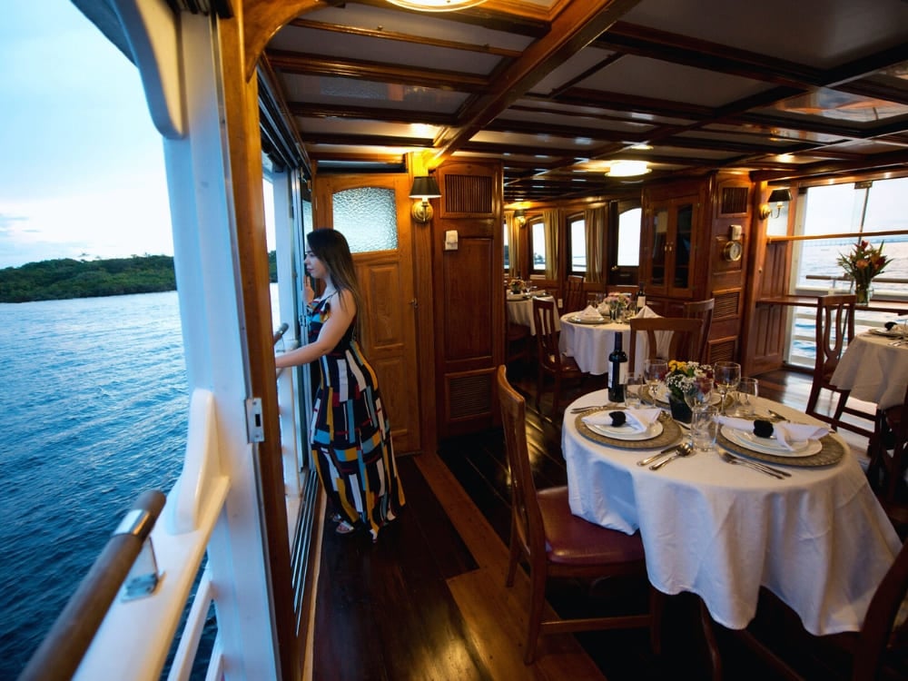 Breathtaking view of a colorful sunset over the Amazon River seen from the bow of the Tucano expedition boat. A woman in a colorful striped dress looks out the open window of a wooden boat dining room, which features tables set with white tablecloths and wine glasses.