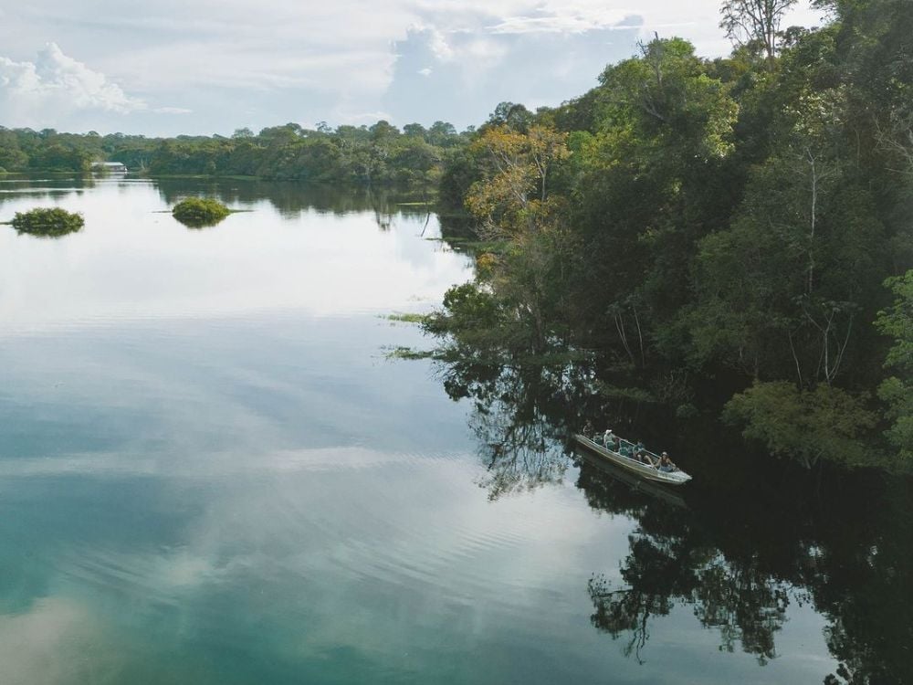 Small boat navigating in the distance on the calm and reflective waters of the Mamori River, surrounded by green jungle vegetation. Small boat navigating in the distance on the calm and reflective waters of the Mamori River, surrounded by green jungle vegetation.