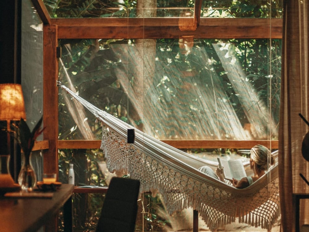 A person relaxing in a hammock inside a sophisticated room with large glass windows facing the jungle. A person relaxing in a hammock inside a sophisticated room with large glass windows facing the jungle.