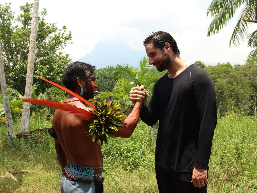 A Yanomami indigenous guide wearing traditional red feathers shaking hands with a hiker in the forest. A Yanomami indigenous guide wearing traditional red feathers shaking hands with a hiker in the forest.