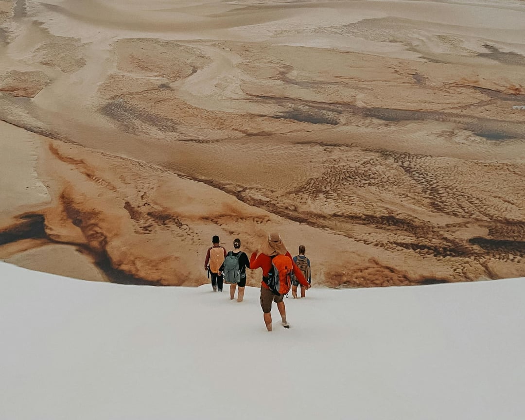 A group of tourists walks across a dry, brown lagoon bed surrounded by large white sand dunes during the dry season. A group of tourists walks across a dry, brown lagoon bed surrounded by large white sand dunes during the dry season.