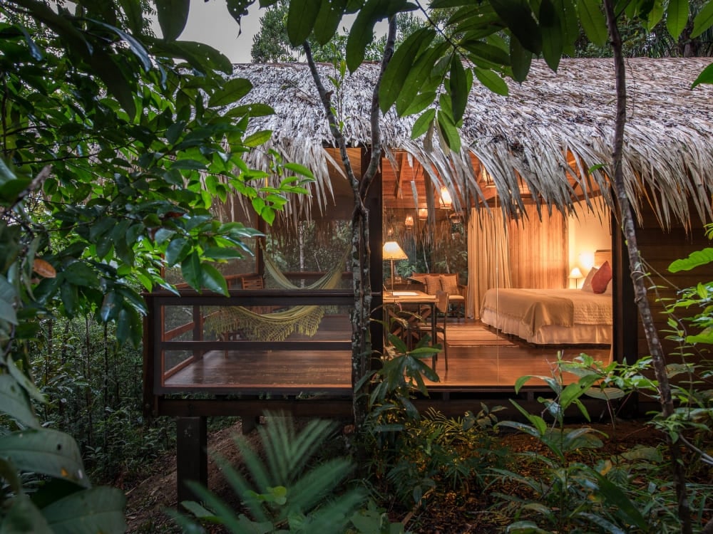 Illuminated rustic luxury bungalow with a thatched roof, comfortable bed, and balcony hammock surrounded by the Brazilian jungle. Illuminated rustic luxury bungalow with a thatched roof, comfortable bed, and balcony hammock surrounded by the Brazilian jungle.
