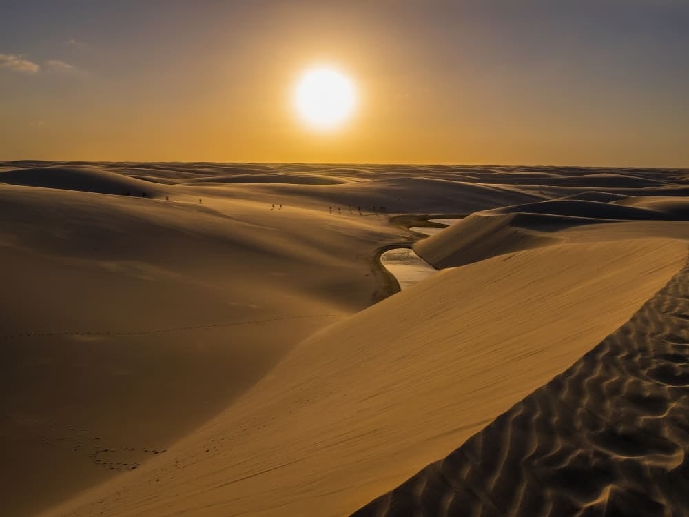 The sun sets brightly over undulating sand dunes and a shallow stream winding through the Lencois Maranhenses National Park in Brazil. The sun sets brightly over undulating sand dunes and a shallow stream winding through the Lencois Maranhenses National Park in Brazil.