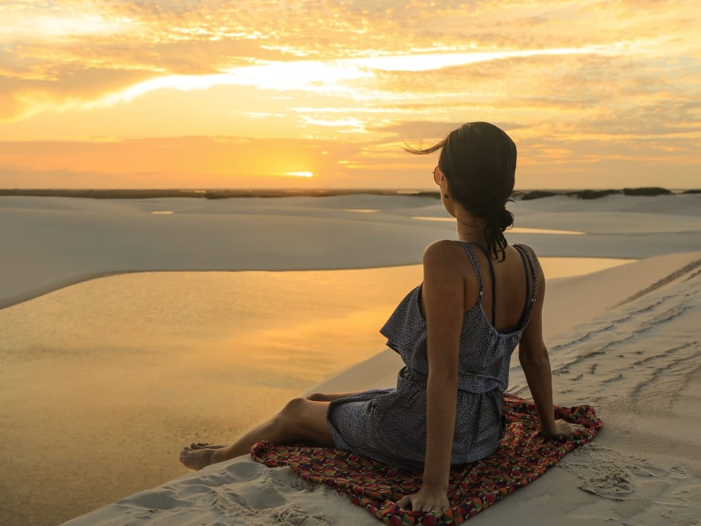 A woman sits on a patterned towel atop a dune, enjoying the warm Lençóis Maranhenses weather and watching the sunset over a serene lagoon. A woman sits on a patterned towel atop a dune, enjoying the warm Lençóis Maranhenses weather and watching the sunset over a serene lagoon.