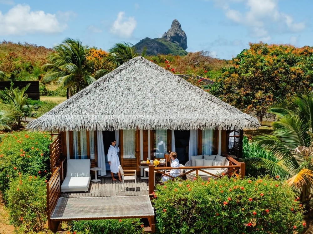 Couple in white bathrobes relaxing on the private deck of a rustic thatched-roof bungalow with views of Morro do Pico at a top hotel in Fernando de Noronha. Couple in white bathrobes relaxing on the private deck of a rustic thatched-roof bungalow with views of Morro do Pico at a top hotel in Fernando de Noronha.