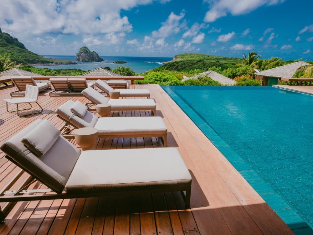 Wooden deck with comfortable lounge chairs beside an infinity pool overlooking the ocean and Morro do Pico at a hotel in Fernando de Noronha Wooden deck with comfortable lounge chairs beside an infinity pool overlooking the ocean and Morro do Pico at a hotel in Fernando de Noronha