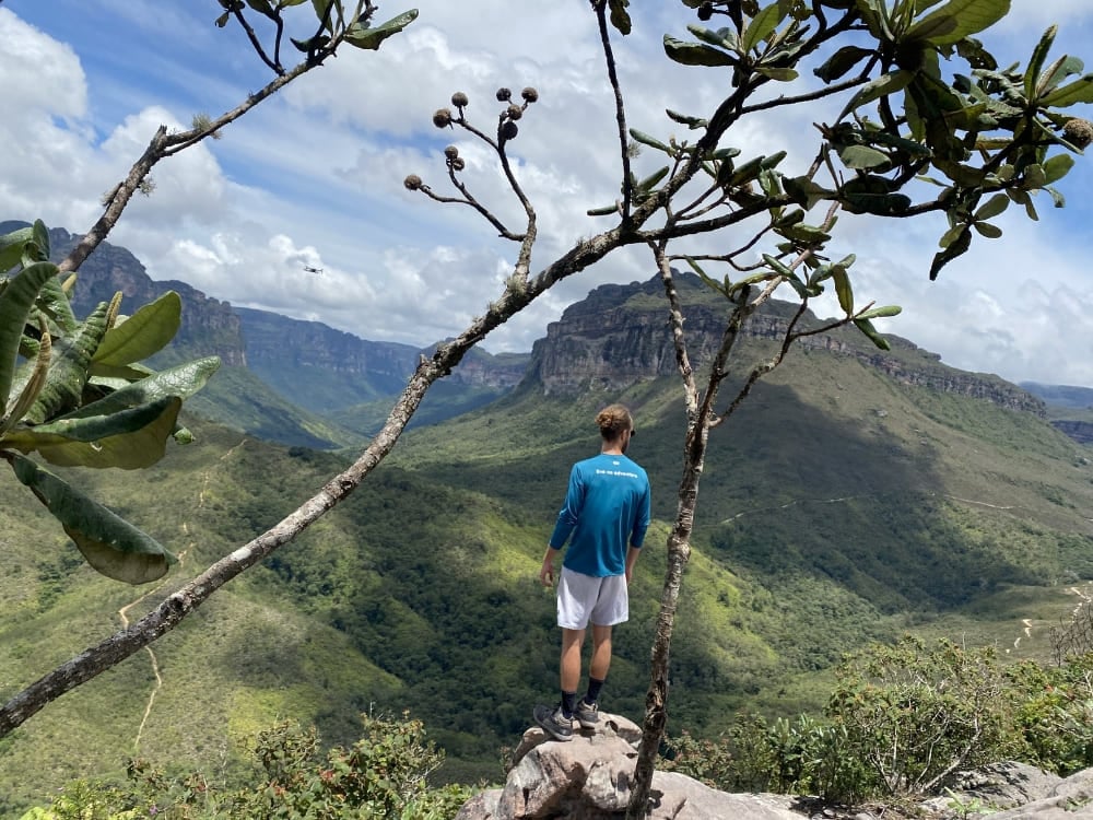 A hiker stands on a rocky edge framed by tree branches, looking out over the green valley toward the flat-topped Morro do Castelo. A hiker stands on a rocky edge framed by tree branches, looking out over the green valley toward the flat-topped Morro do Castelo.