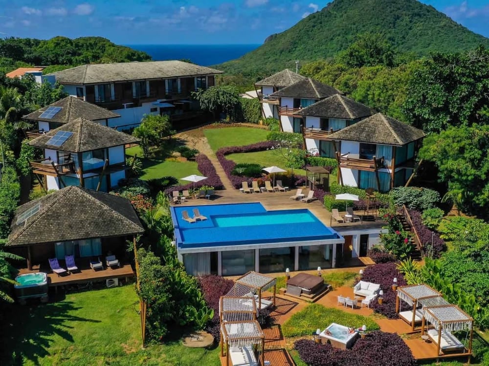 Aerial view of Pousada Morena featuring dark-roofed cabins, a central blue swimming pool, and hot tubs surrounded by lush forests in Fernando de Noronha. Aerial view of Pousada Morena featuring dark-roofed cabins, a central blue swimming pool, and hot tubs surrounded by lush forests in Fernando de Noronha.
