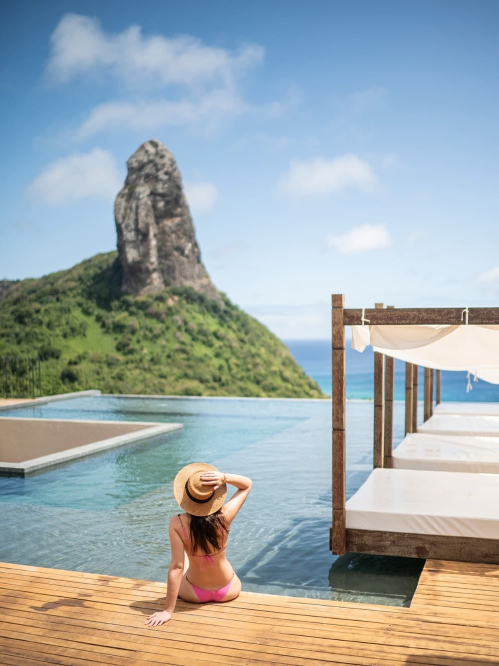 Woman wearing a straw hat relaxing on a wooden deck by an infinity pool with a stunning view of Morro do Pico at a luxury hotel in Fernando de Noronha, Brazil. Woman wearing a straw hat relaxing on a wooden deck by an infinity pool with a stunning view of Morro do Pico at a luxury hotel in Fernando de Noronha, Brazil.