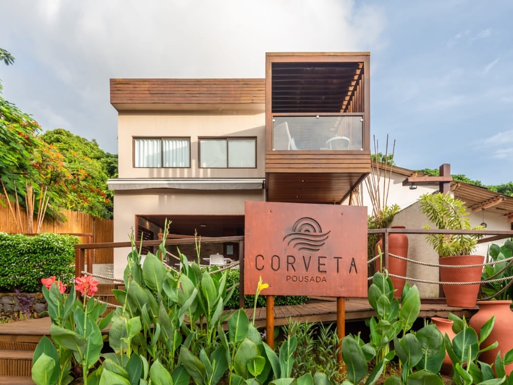 Modern exterior of Pousada Corveta with a metal sign, wooden walkways, and tropical plants, highlighting where to stay in Fernando de Noronha. Modern exterior of Pousada Corveta with a metal sign, wooden walkways, and tropical plants, highlighting where to stay in Fernando de Noronha.