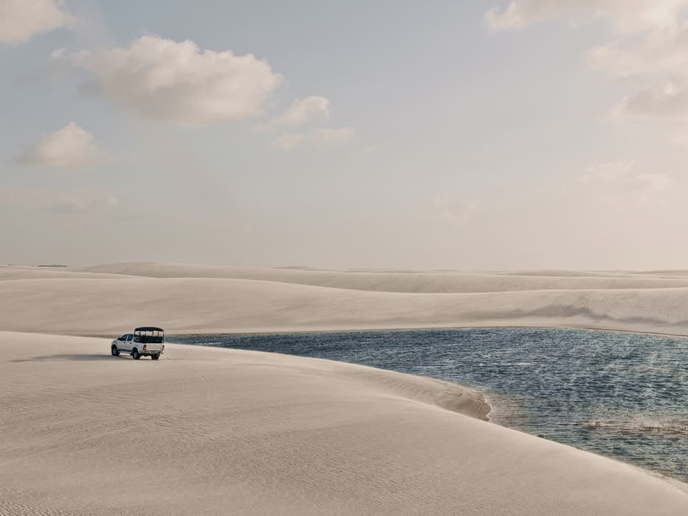 A white 4x4 tour vehicle driving across vast white sand dunes next to a bright blue lagoon. A white 4x4 tour vehicle driving across vast white sand dunes next to a bright blue lagoon.