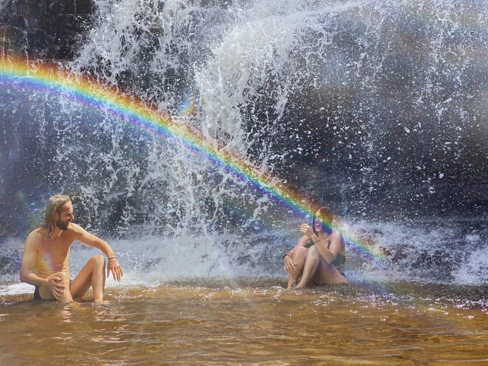 Two travelers relax in the shallow golden waters of a Chapada Diamantina waterfall pool, with a vivid rainbow shining across the mist. Two travelers relax in the shallow golden waters of a Chapada Diamantina waterfall pool, with a vivid rainbow shining across the mist.