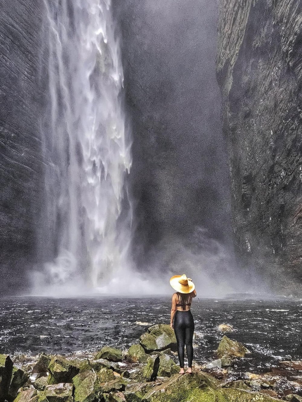 A hiker wearing a hat stands at the edge of a dark pool, looking up at the dramatic 100-meter vertical drop of Fumacinha Waterfall inside a narrow canyon. A hiker wearing a hat stands at the edge of a dark pool, looking up at the dramatic 100-meter vertical drop of Fumacinha Waterfall inside a narrow canyon.