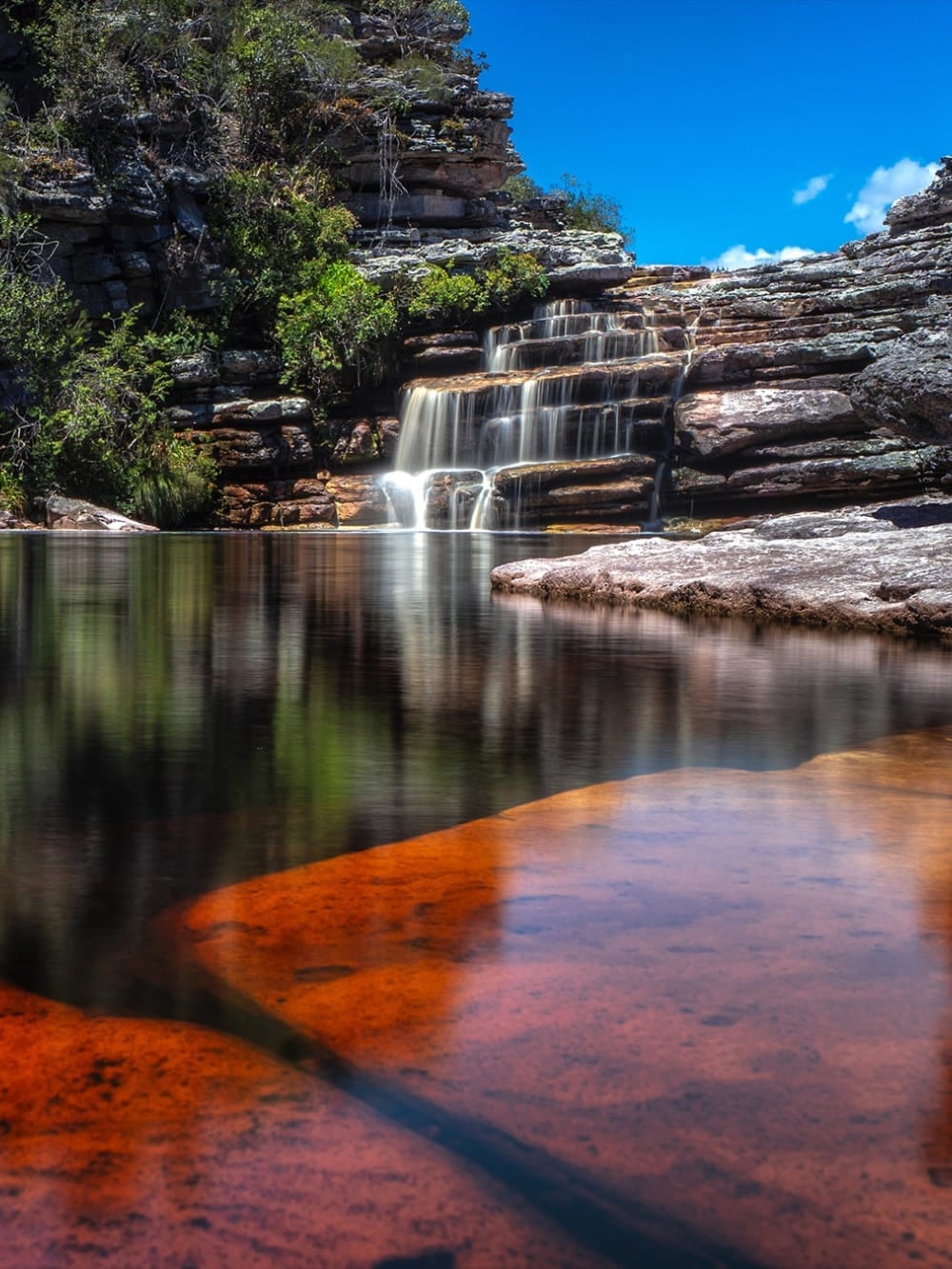 A cascading waterfall flowing into a calm natural pool with red rocks in Chapada Diamantina National Park. A cascading waterfall flowing into a calm natural pool with red rocks in Chapada Diamantina National Park.