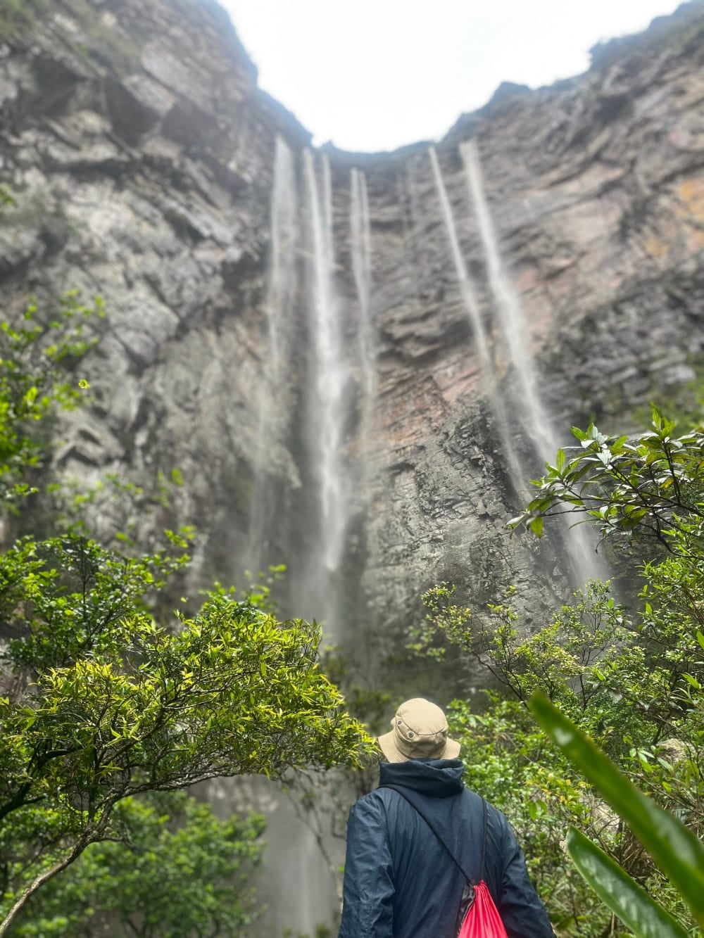 A hiker wearing a hat and jacket looks up at the towering 280-meter water streams of Cachoeirão Waterfall from its rocky base. A hiker wearing a hat and jacket looks up at the towering 280-meter water streams of Cachoeirão Waterfall from its rocky base.