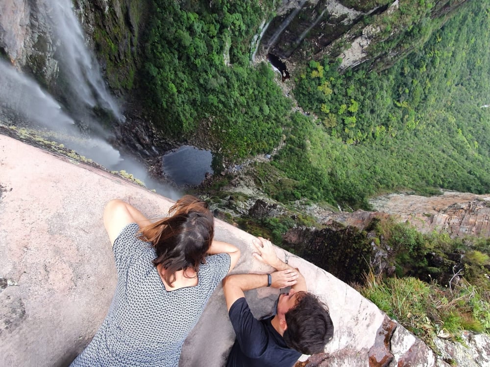 Two hikers look down from a high cliff edge at Cachoeirão Waterfall plunging into the deep green Vale do Pati canyon. Two hikers look down from a high cliff edge at Cachoeirão Waterfall plunging into the deep green Vale do Pati canyon.