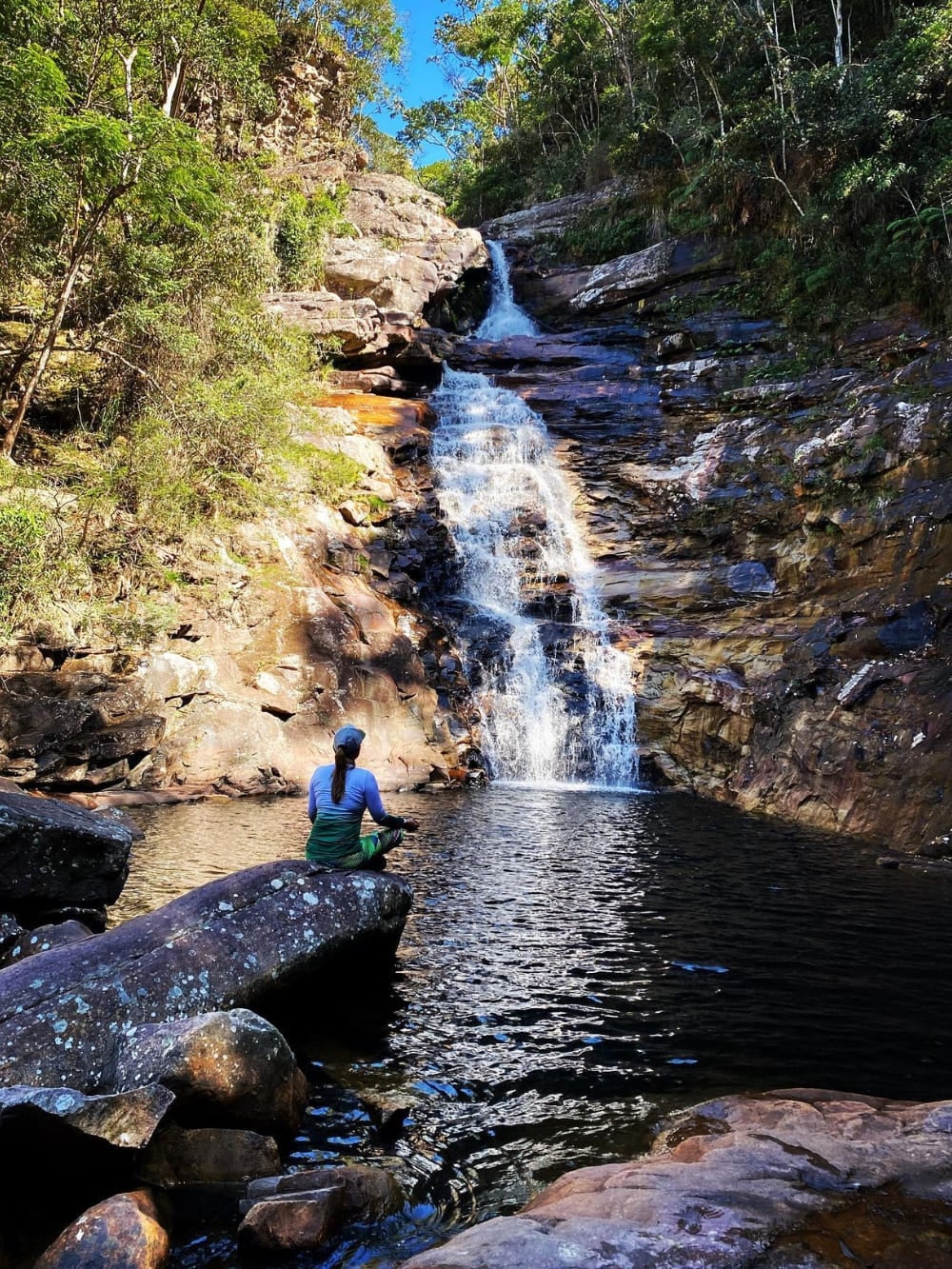 A woman sits on a rock near the calm natural pool of Funil Waterfall, a 20-meter drop surrounded by dense green vegetation. A woman sits on a rock near the calm natural pool of Funil Waterfall, a 20-meter drop surrounded by dense green vegetation.