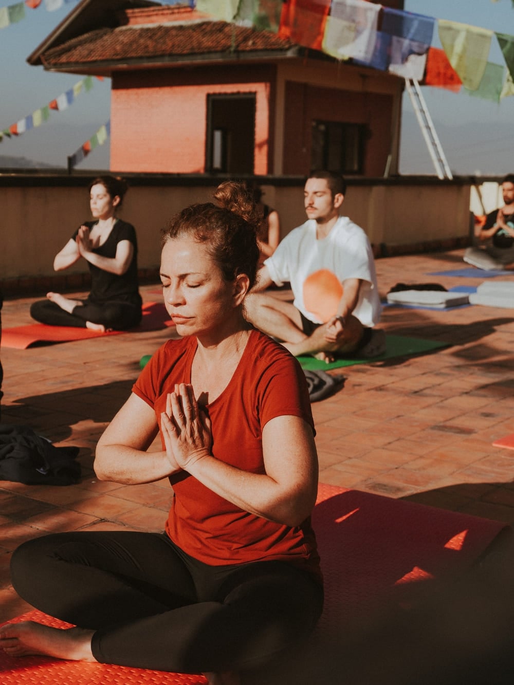 Mila Ricetti and Lorenzo practicing yoga and meditating with a group on a sunlit rooftop decorated with prayer flags during a retreat in Nepal. Mila Ricetti and Lorenzo practicing yoga and meditating with a group on a sunlit rooftop decorated with prayer flags during a retreat in Nepal.