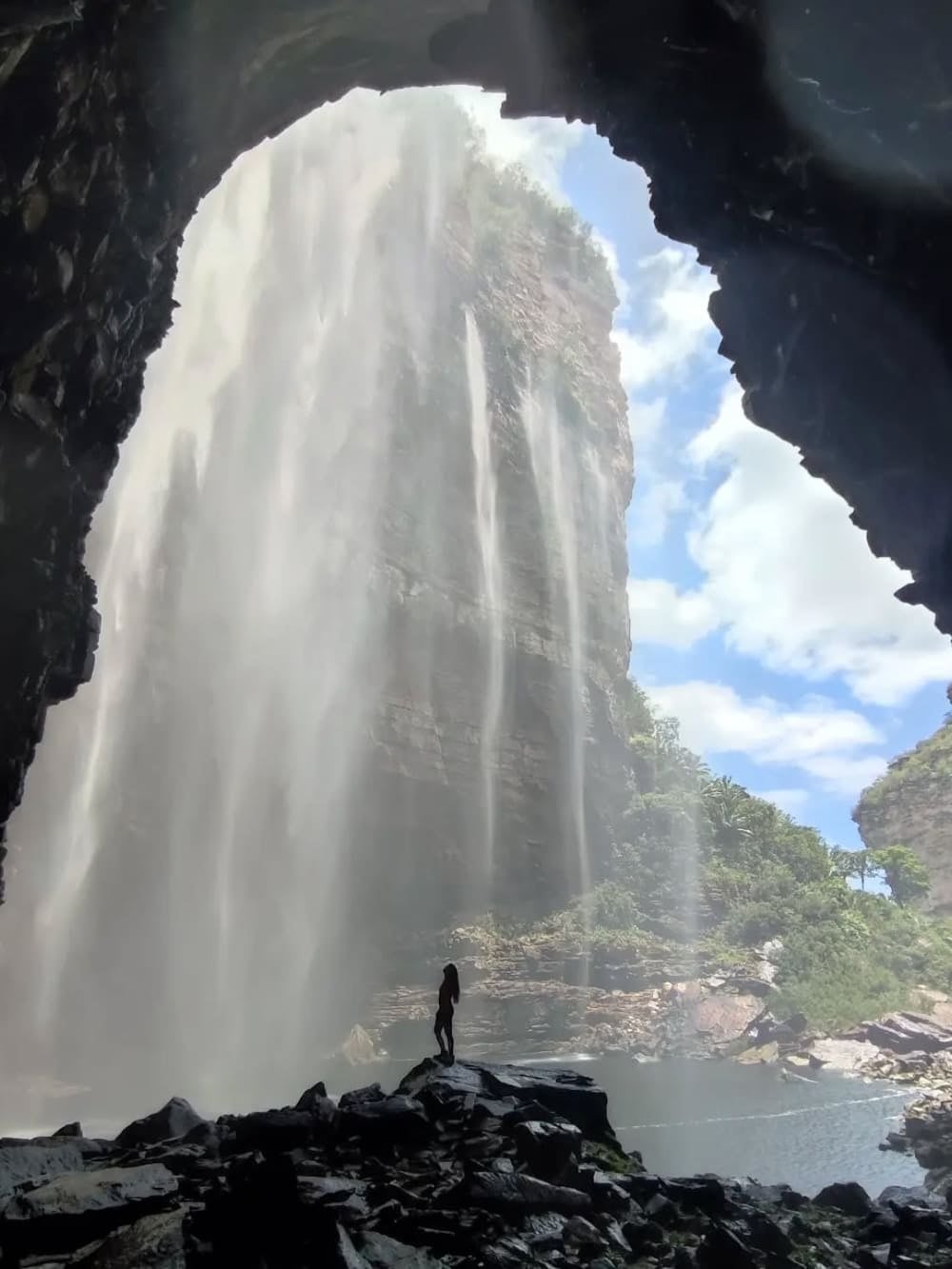 A silhouette of a person standing on rocks near a dark cave entrance, looking out at the rugged 100-meter cliffs of Herculano Waterfall. A silhouette of a person standing on rocks near a dark cave entrance, looking out at the rugged 100-meter cliffs of Herculano Waterfall.