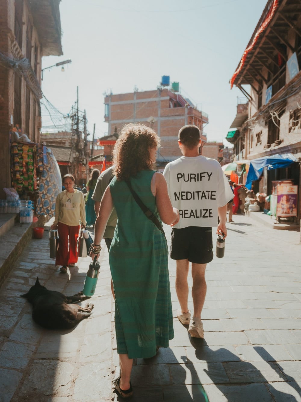 Mila and Lorenzo walking side by side down a sunny street in Nepal, with Lorenzo wearing a t-shirt that says purify, meditate, realize. Mila and Lorenzo walking side by side down a sunny street in Nepal, with Lorenzo wearing a t-shirt that says purify, meditate, realize.