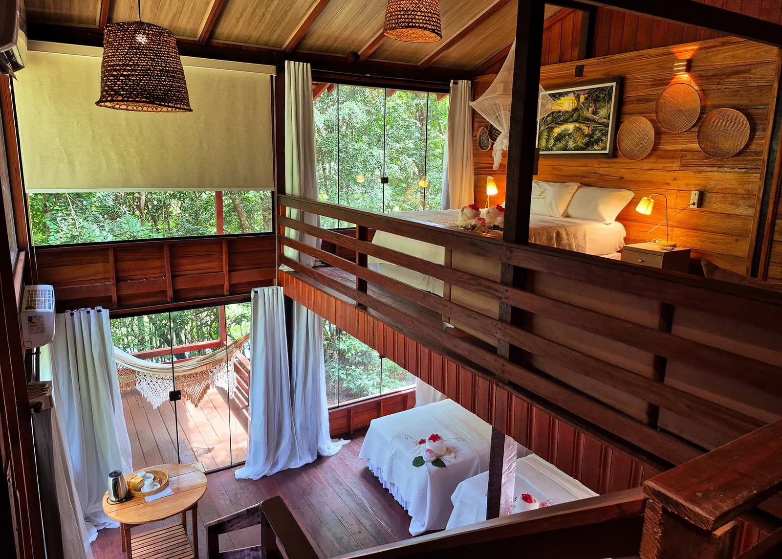 Your own corner of the forest, with rose petals and a hammock waiting. Interior view from the mezzanine of an Amazon boutique lodge bungalow, showing a loft bedroom with warm wooden walls, white linens, wicker pendant lights, and floor-to-ceiling windows overlooking dense green forest, with a lower living area featuring a massage table decorated with rose petals, sheer white curtains, and a hammock on the deck outside