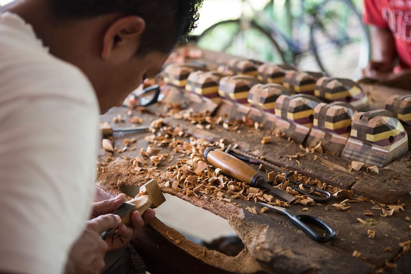 Every curl of wood on that bench is a decision, not an accident. A craftsman carves a small wooden piece by hand on a workbench scattered with wood shavings, with rows of multicoloured laminated wood figurines displayed behind him — a local artisan encounter on an Amazon jungle cruise Brazil