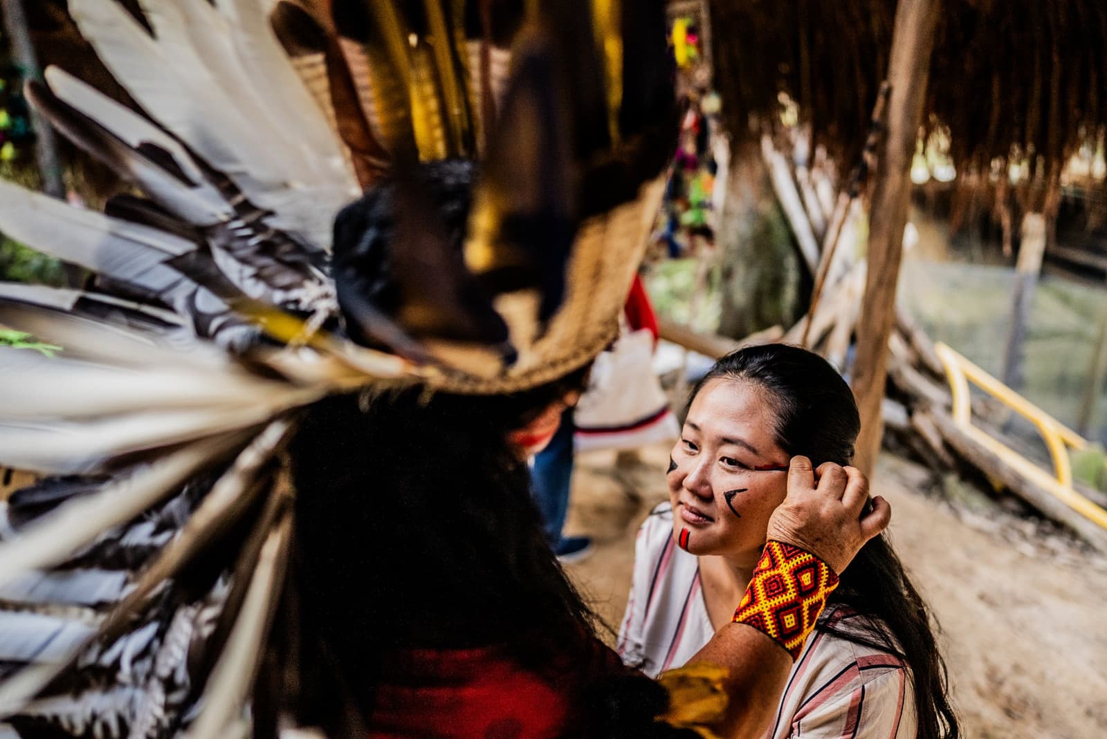 Face paint, a feather headdress, and a smile that says this moment is real. Photo: FG Trade A smiling visitor with traditional geometric face paint and a beaded wrist cuff sits face-to-face with an Indigenous community member wearing a large feather headdress during an amazon river cruise brazil cultural visit