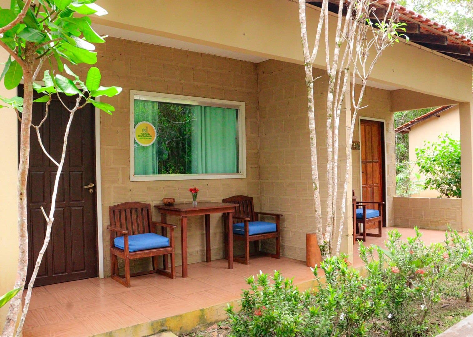 Your own little porch, just steps from the forest. A shaded veranda at Amazon Turtle Lodge with two wooden chairs and blue cushions beside a small table, set among tropical garden shrubs and slender trees against a stone-brick bungalow wall