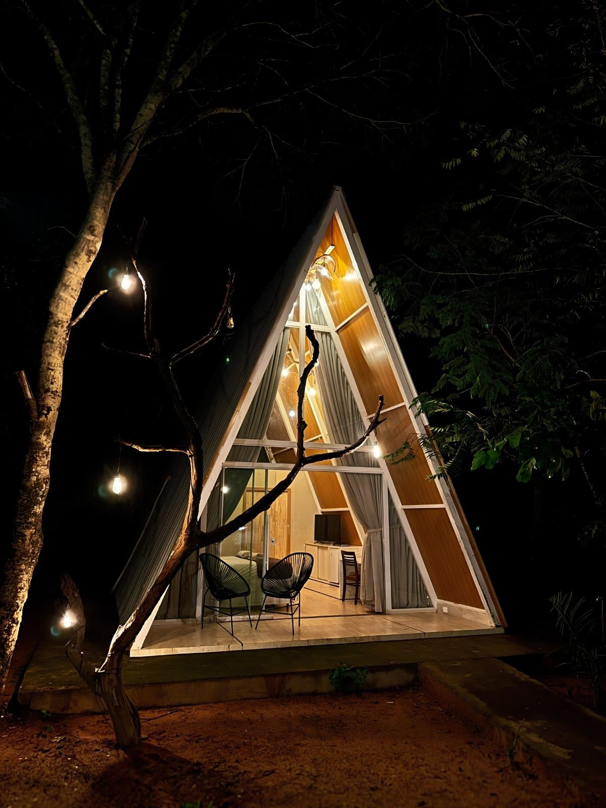 Your shelter in the Cerrado knows how to set the mood. A illuminated A-frame cabin glowing with warm light at night, surrounded by dark trees and strung with Edison bulbs, with two chairs on the open deck — a cozy base for a Jalapão adventure
