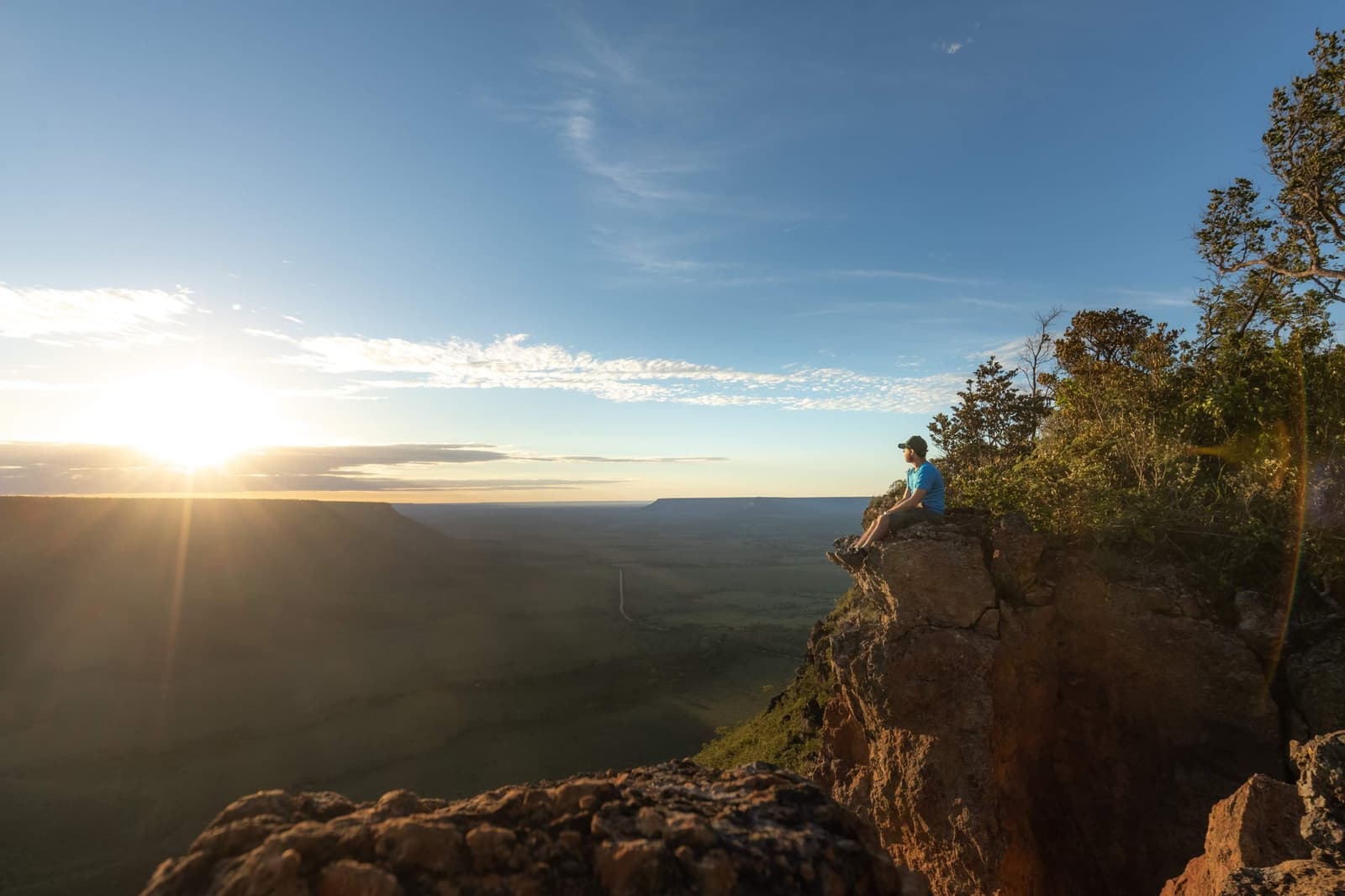 Up before dawn, rewarded with the whole park at your feet. Photo: Alexandre Socci A traveler sits on a rocky cliff edge at sunrise on a Jalapão adventure, gazing over a vast Cerrado plateau with golden light fanning across the horizon and a distant mesa in view