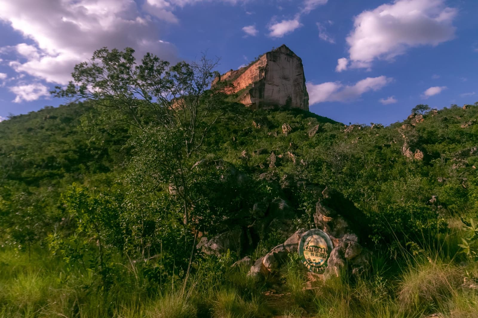 The trail sign is just the beginning, the rock does the real talking. Photo: Alexandre Socci Low-angle view up a lush green hillside toward a large flat-topped sandstone rock formation, with a Catedral do Jalapão trail marker sign on a boulder in the foreground under a blue sky with scattered clouds