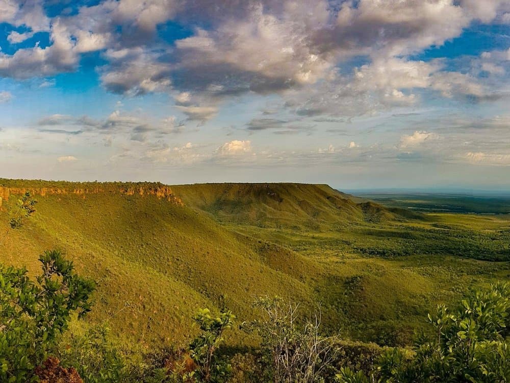 Four hundred metres of open sky, and the cerrado laid out like it has nowhere to be. Panoramic view from the top of a sandstone plateau during a Jalapão trekking route, showing green cerrado-covered slopes, red cliff faces, and an expansive lowland stretching to the horizon under a wide sky with scattered clouds