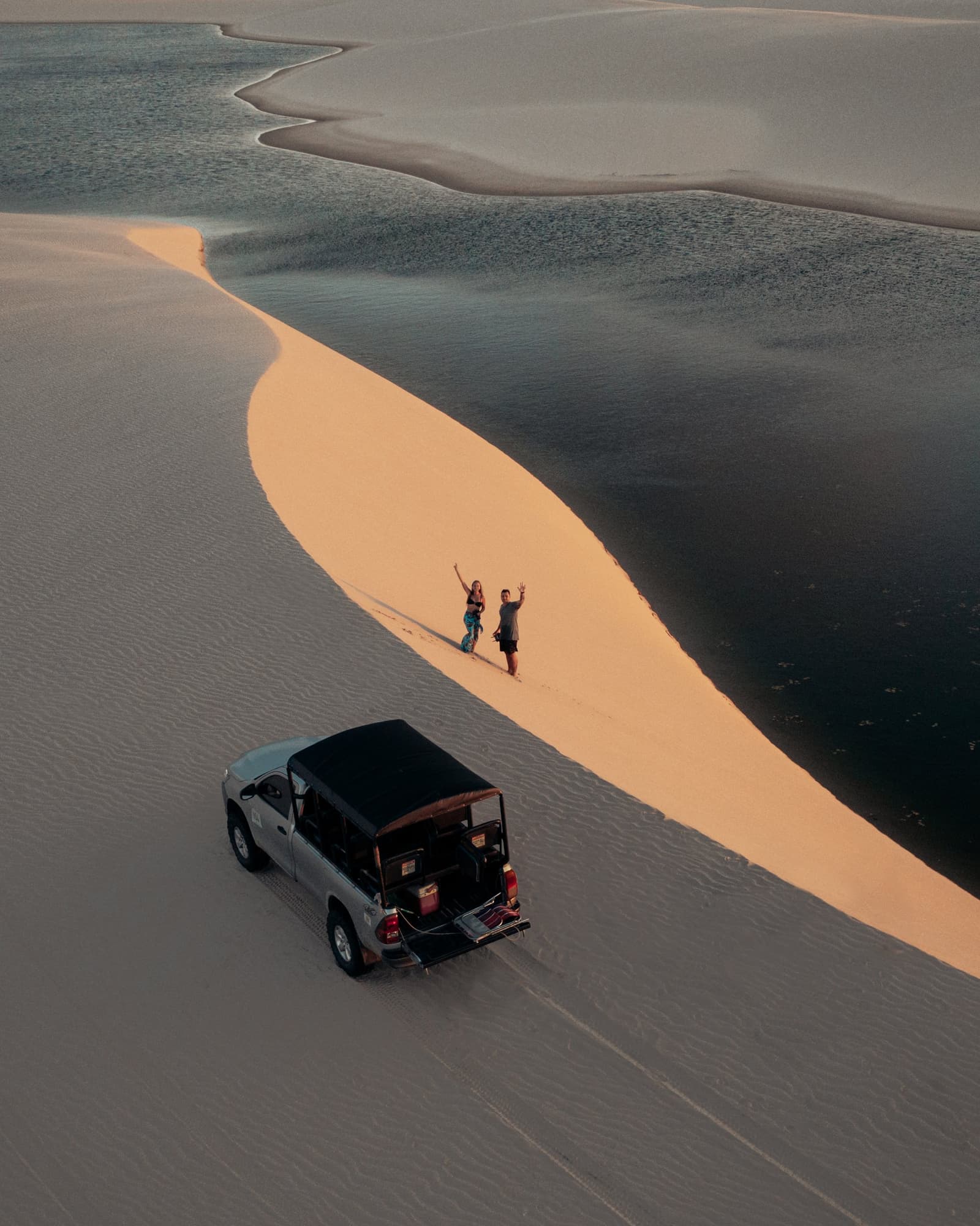 Two people, one dune ridge, zero signal, and they couldn't be happier. Aerial view of two travelers waving from a golden sand dune ridge beside a dark reflective lagoon during sunset, with a 4x4 vehicle parked below — a scene from a Lençóis Maranhenses full itinerary