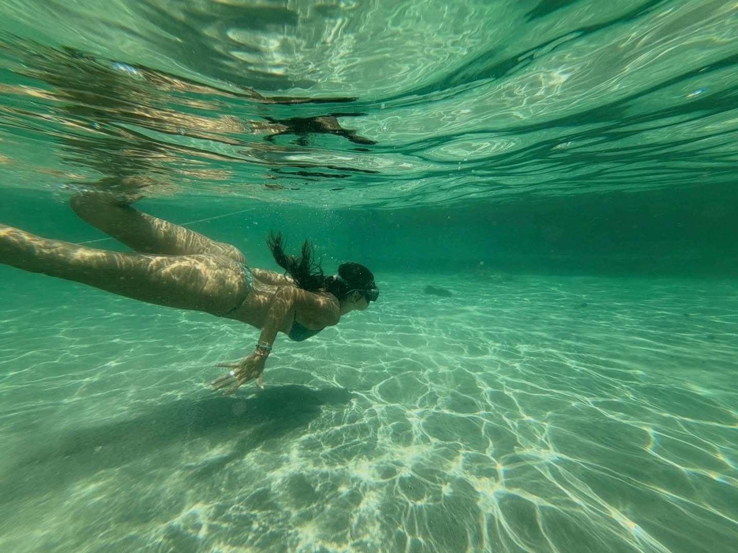 Turns out the best view of the lagoon is from inside it. A woman free-diving underwater through clear green-tinted shallows during a Lençóis Maranhenses hike, sunlight dappling the sandy floor below her