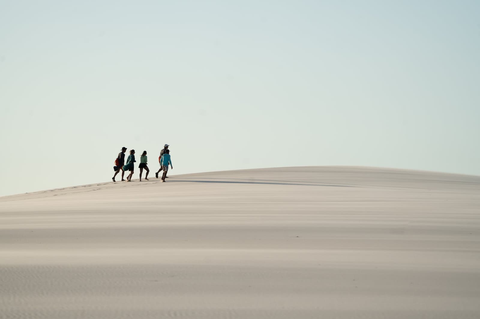 Four tiny figures, one enormous dune, scale does the talking. Four trekkers walking along the curved crest of a vast white sand dune during a Lençóis Maranhenses hike, under a pale blue sky