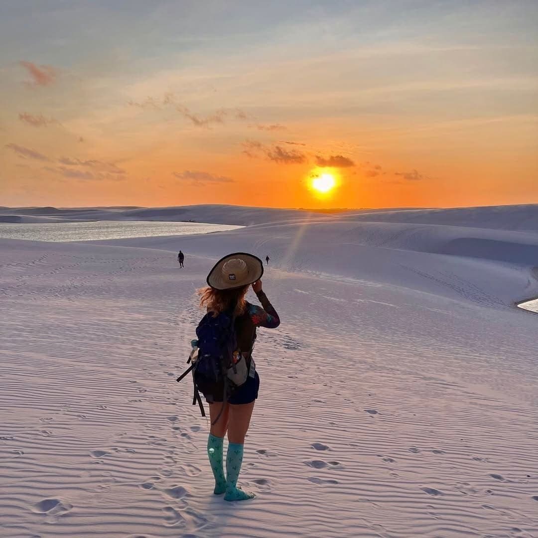 Hat in hand, sun going down, the dunes have a way of slowing you right up. A hiker with a wide-brim hat and backpack stands barefoot in turquoise socks on white sand dunes during the Lençóis Maranhenses hike, watching an orange sunset over distant lagoons and scattered figures on the dunes