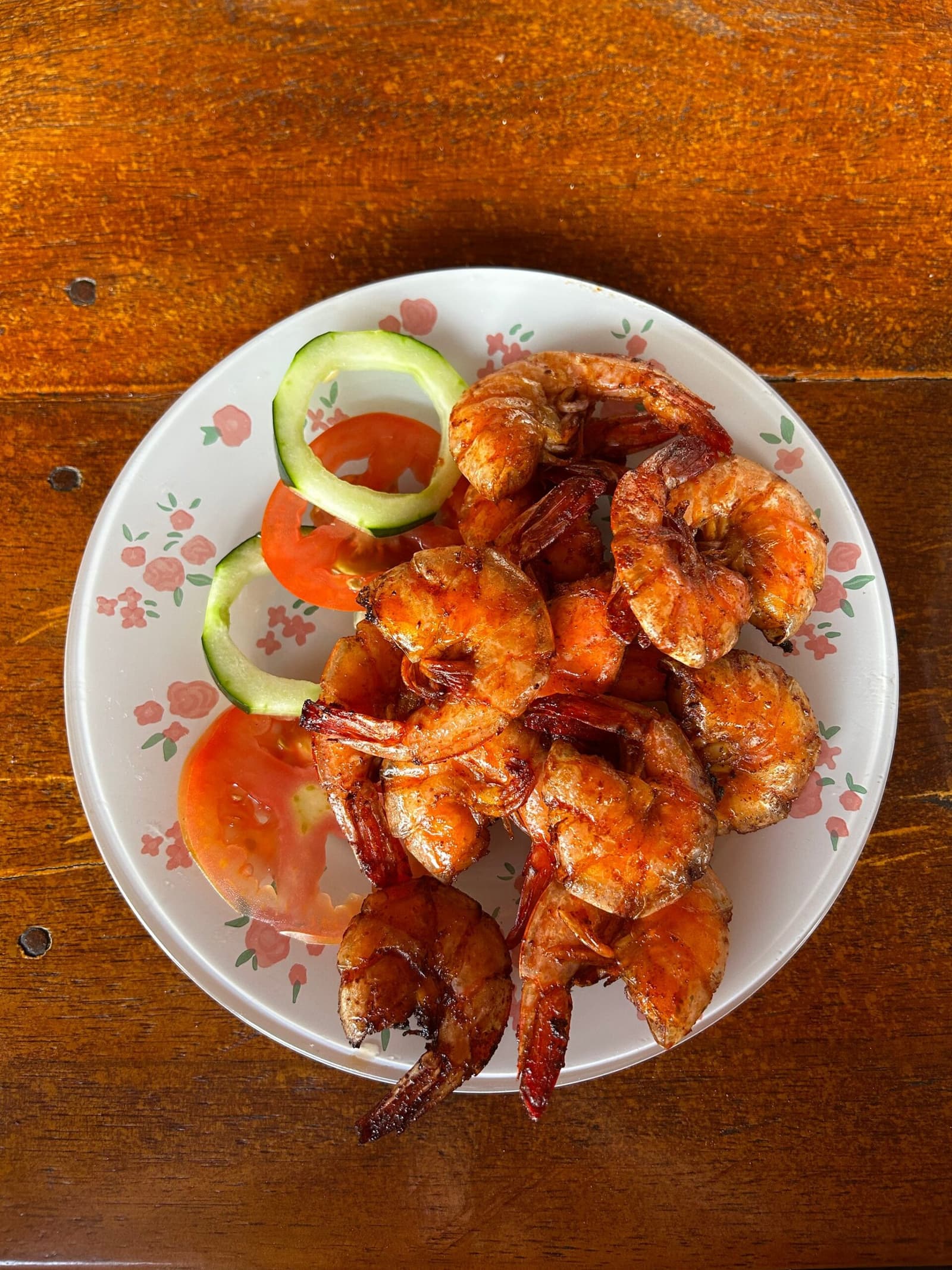 Fresh from the coast and straight to the table, no ceremony needed. A plate of grilled whole shrimp with a golden-red glaze, garnished with sliced tomato and cucumber rings, served on a floral-rimmed plate on a wooden table during a lencois maranhenses trek meal