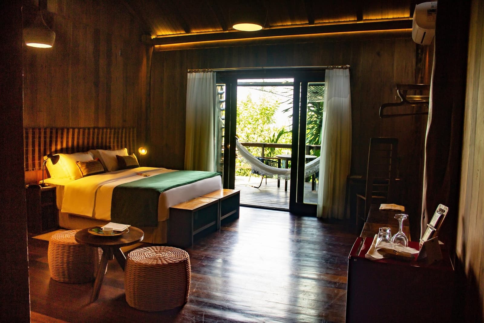 The hammock is right there, no excuses for not napping in the jungle. A warmly lit timber cabin bedroom at Mirante do Gavião amazon lodge, with a double bed dressed in yellow and green linens, wicker ottomans, and open French doors leading to a private balcony with a hammock overlooking lush forest canopy