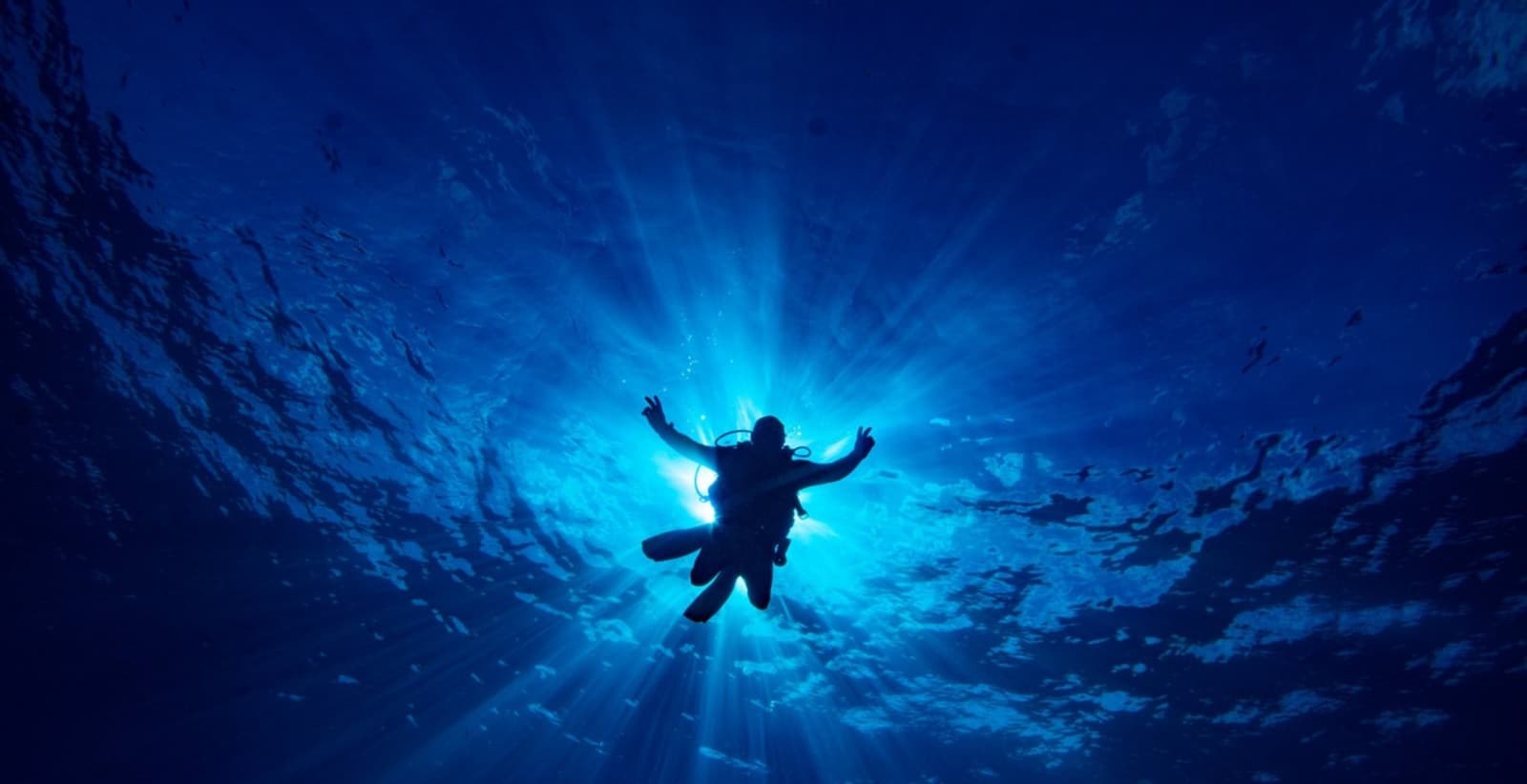 Arms wide open, forty feet down. Good answer to almost any question. Silhouette of a scuba diver floating in deep blue water with arms raised, backlit by rays of sunlight during an Open Water diving course