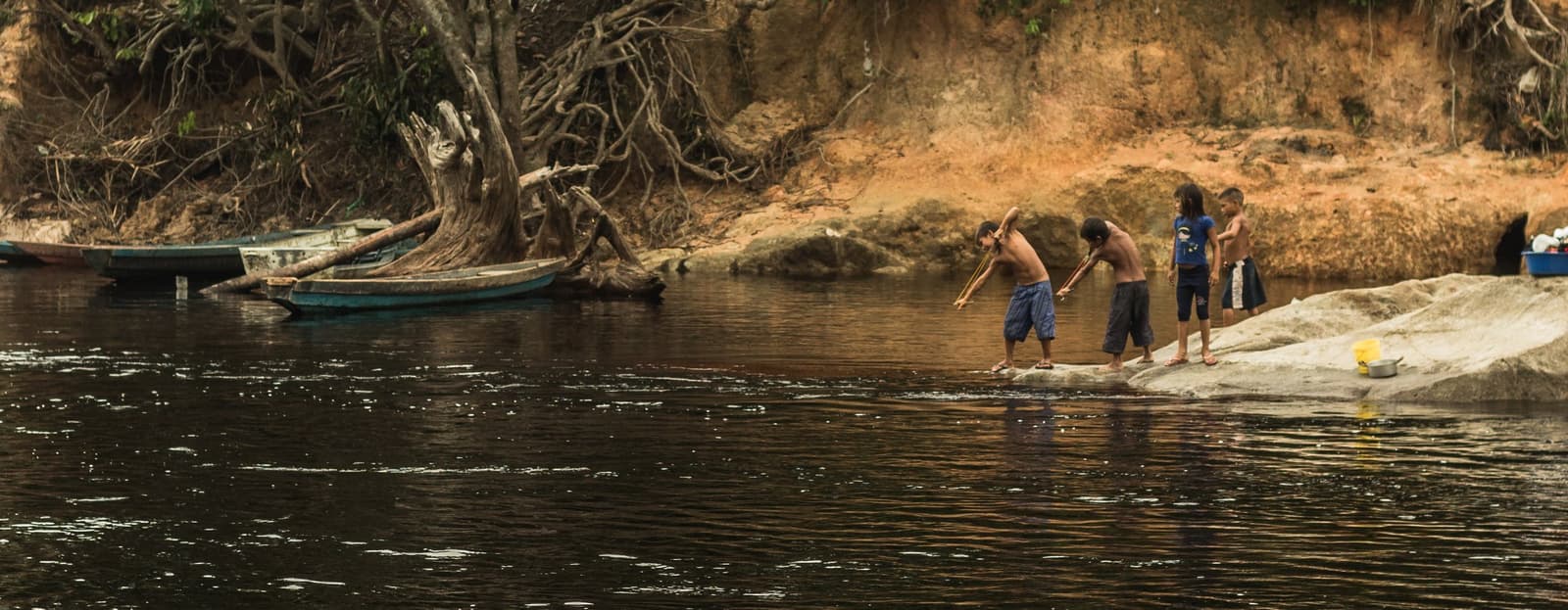 River life runs on its own schedule, and kids here learned it long before any visitor arrived. Four children stand on a flat rock at the edge of a dark Amazon river, using long sticks to fish or play, with wooden canoes and exposed tree roots along a sandy clay bank behind them — a riverside scene encountered during the Pico da Neblina Expedition